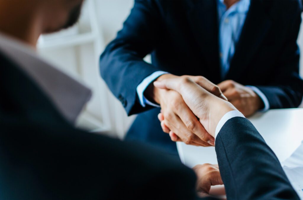 Dois homens em trajes formais apertando as mãos em um ambiente corporativo, simbolizando uma parceria de negócios e confiança mútua. 