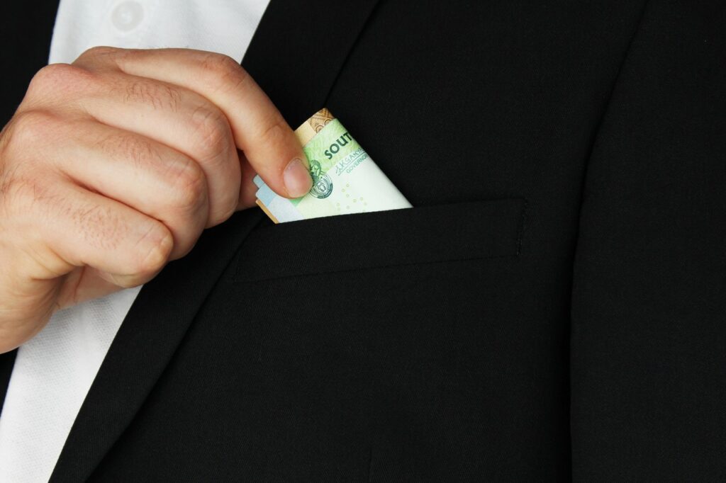 Um homem vestindo um terno preto coloca discretamente uma nota de dinheiro no bolso do paletó, simbolizando corrupção, suborno ou práticas financeiras ilícitas.






