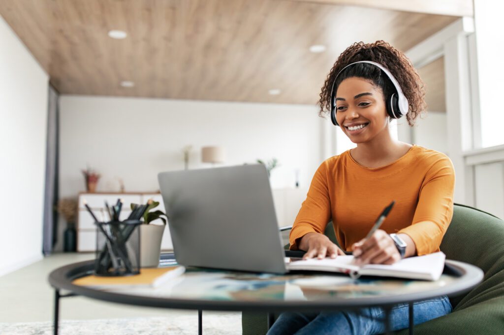 Mulher sorridente, usando fones de ouvido e blusa laranja, trabalha em um laptop enquanto faz anotações em um caderno. Ela está sentada em uma sala bem iluminada, com móveis modernos e decoração minimalista ao fundo.