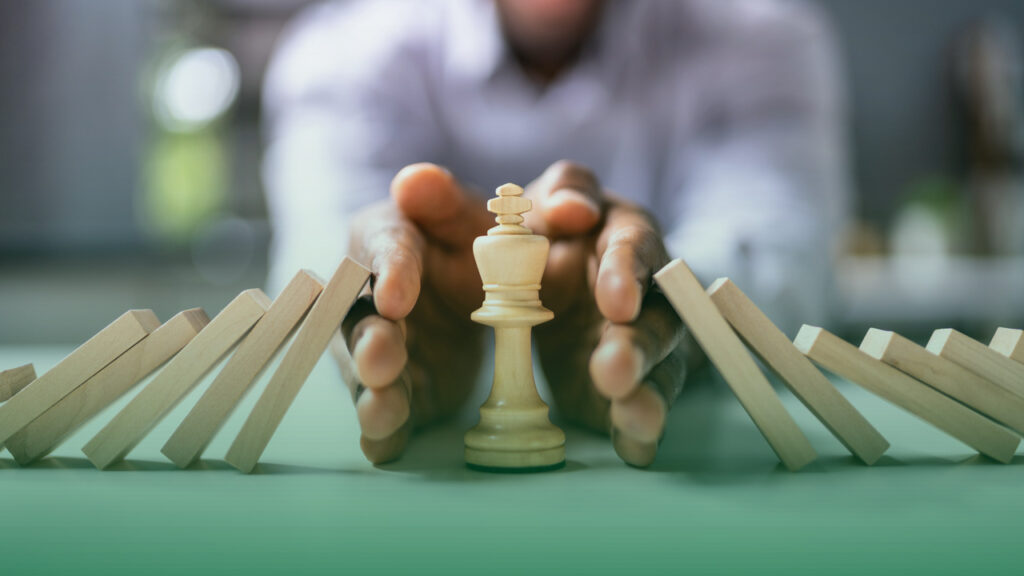 Mão de uma pessoa interrompendo o efeito dominó de blocos de madeira que estão caindo, protegendo uma peça de xadrez (um rei) no centro. A imagem representa a ideia de proteção e controle de riscos, simbolizando ações para evitar consequências negativas em uma sequência de eventos.