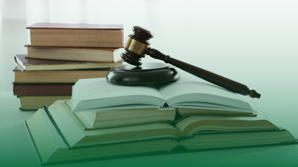 Um martelo de juiz repousando sobre um livro aberto, com pilhas de livros jurídicos ao fundo, representando a conexão entre legislação e compliance