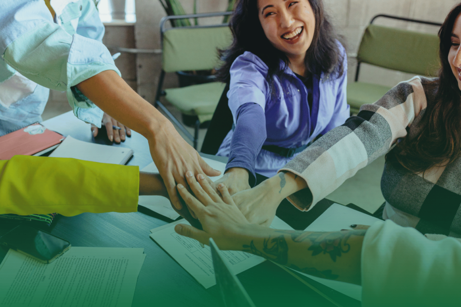 Grupo de colaboradores com mãos unidas sobre a mesa, sorrindo, simbolizando união, diversidade e colaboração no ambiente corporativo.
