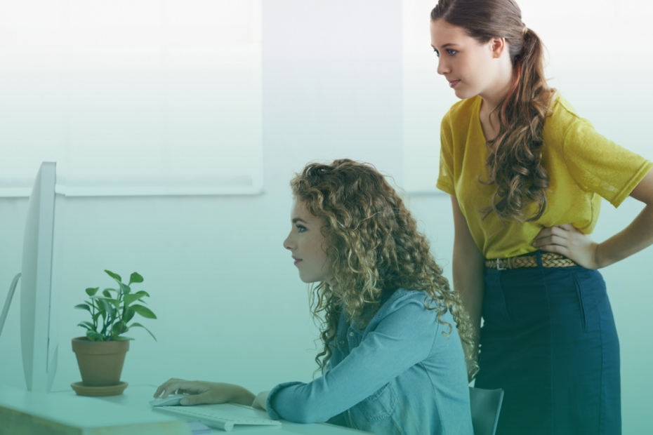 Duas mulheres analisam informações em um computador de mesa em ambiente corporativo, transmitindo foco e atenção ao realizar uma pesquisa ou verificação de dados.