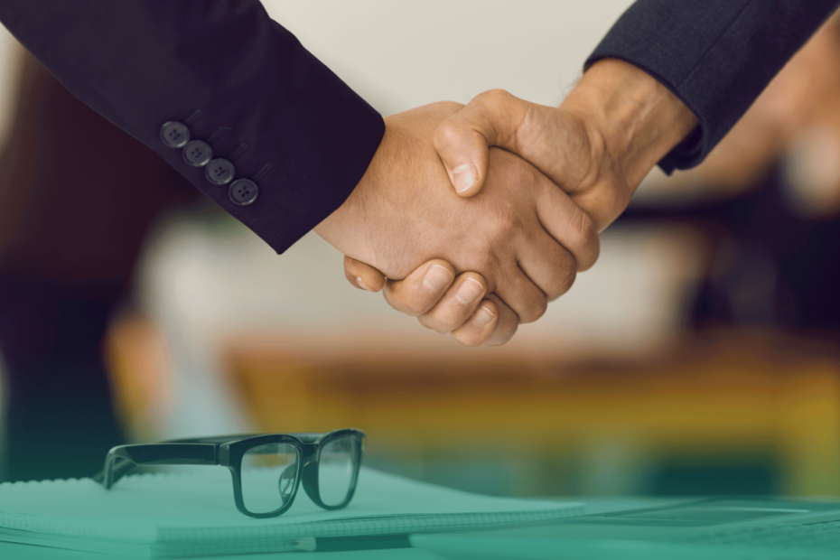 Duas pessoas apertando as mãos em um ambiente corporativo, simbolizando a formalização de uma contratação. Em primeiro plano, um caderno e um par de óculos sobre a mesa.